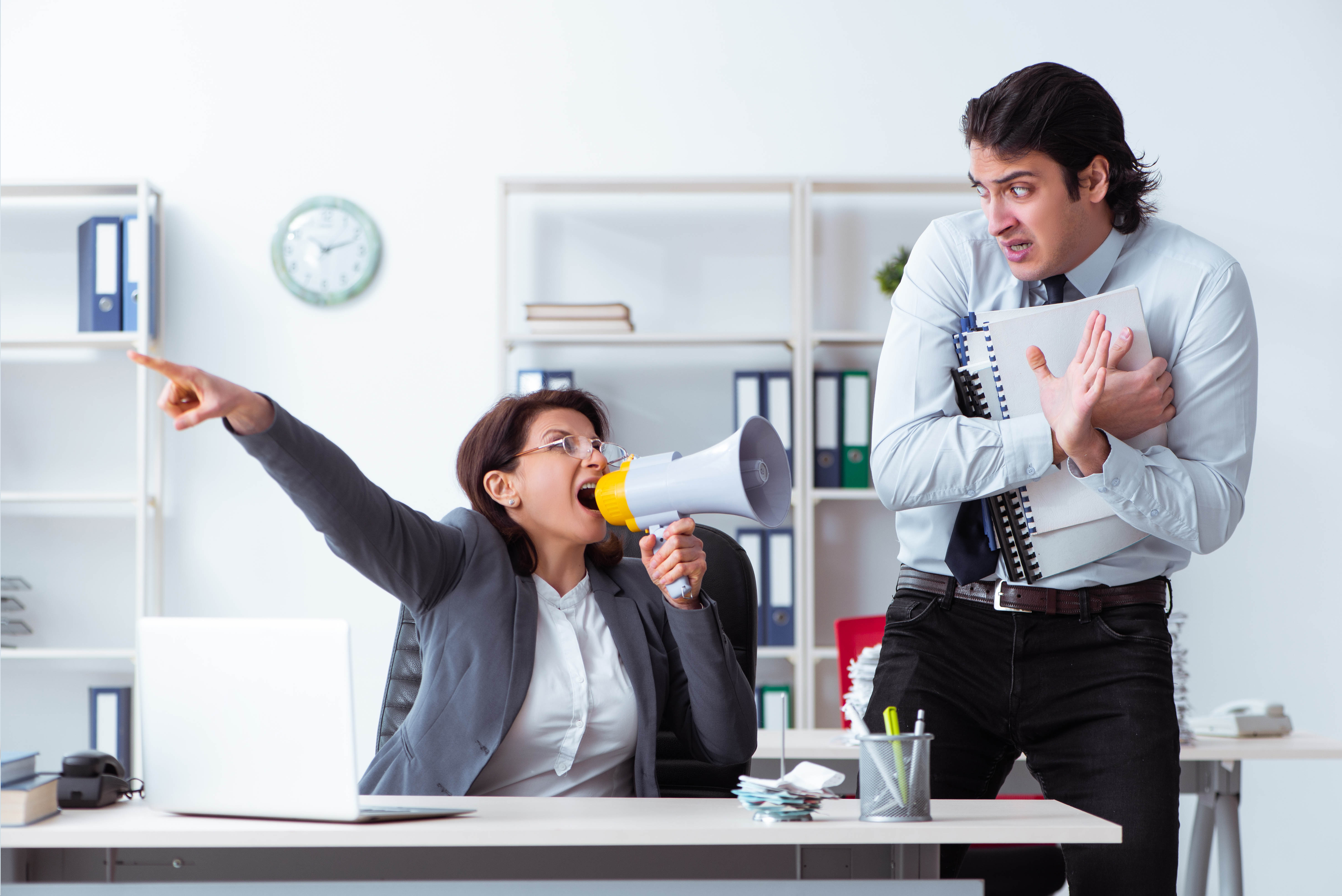 Woman yelling through a megaphone at a young male subordinate who is cowering away Woman yelling through a megaphone at a young male subordinate who is cowering away